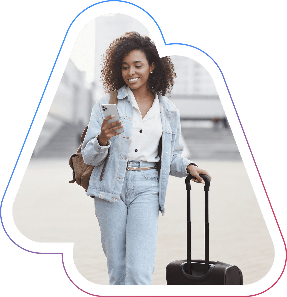 Smiling woman with curly hair in denim outfit, checking her phone while pulling a suitcase outdoors.